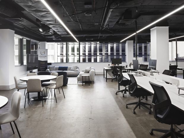 A bright open-plan contemporary office interior with exposed dark services and ductwork above, linear LED strip lighting, polished concrete flooring, white structural columns, extensive glazing, shared workstations with black office chairs, and a lounge and meeting area with sofas and round tables.
