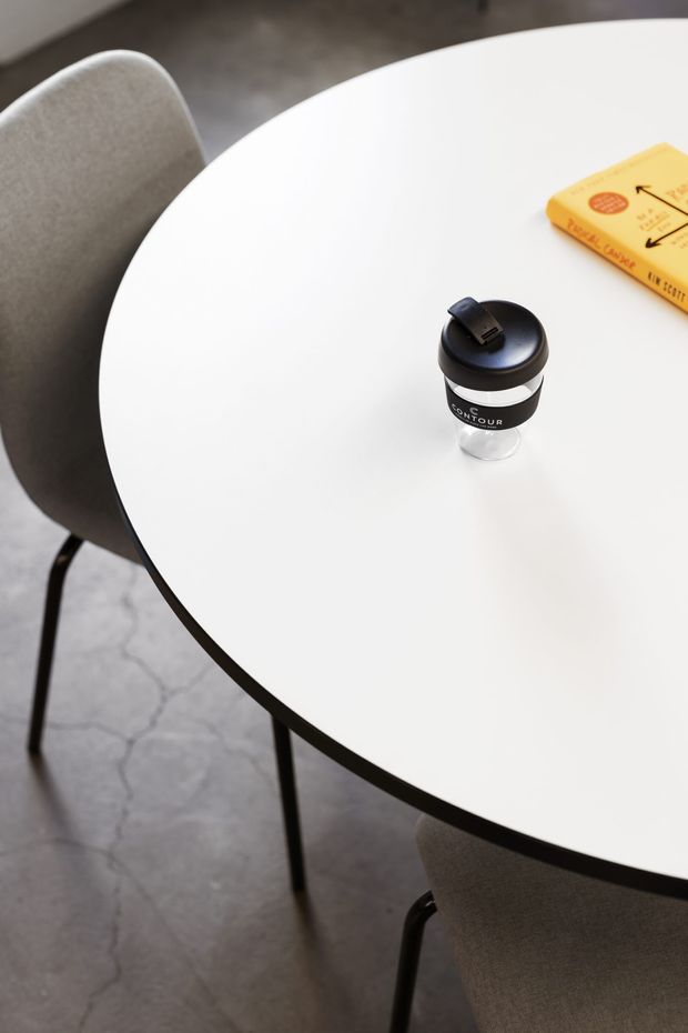 Close-up view of a minimalist round white dining or café table with a slim dark edge and black legs, surrounded by upholstered grey chairs. A reusable coffee cup and yellow book sit on the tabletop. The setting has a contemporary, neutral-toned interior with a grey polished concrete floor.