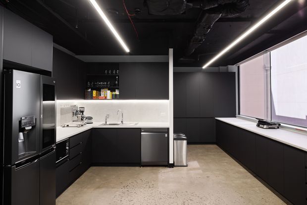 A sleek, contemporary commercial-style kitchen or kitchenette with matte black cabinetry, integrated appliances, a pale stone or solid-surface benchtop, and a softly illuminated light splashback. The space has an exposed black ceiling with linear LED strip lighting, polished concrete flooring, and large windows along one side.