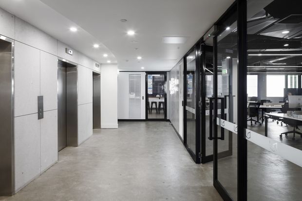A modern commercial office corridor with polished concrete flooring, pale wall panels, recessed ceiling lighting, stainless-steel elevator doors, and black-framed glazed office partitions. Workstations and office furniture are visible through the glass on the right.