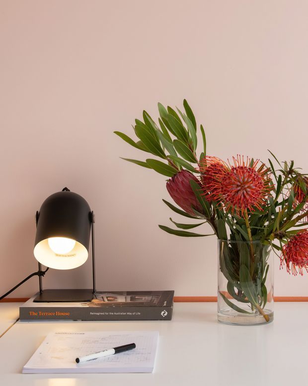 A styled tabletop vignette featuring a black dome desk lamp, an open notebook with pen, an architectural book, and a clear glass vase filled with green foliage and vivid red-orange flowers. The arrangement sits against a warm blush-pink wall with soft, refined lighting.