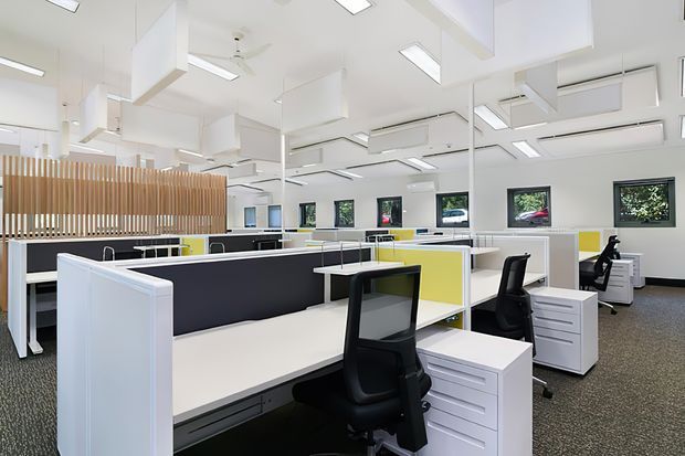 Bright open-plan commercial office interior with rows of white workstations, dark privacy panels, yellow accent partitions, black ergonomic office chairs, filing pedestals, and large windows. The space has a white ceiling with extensive recessed linear lighting, suspended acoustic panels, ceiling fans, and air-conditioning units. A timber vertical-slat divider partially screens the rear area.