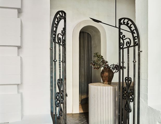 An ornate entryway framed by open black wrought-iron gates with scrolling decorative detailing. White rendered masonry walls and a rounded archway lead to a dark timber-lined passage, while a fluted light-coloured pedestal with a ceramic vase and branches forms a central feature. Dark tiled flooring and restrained neutral finishes create a refined, old-world atmosphere.