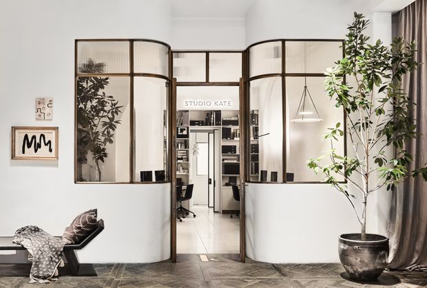 A refined interior view toward a home office or studio enclosed by curved, fluted-glass and dark-framed partitions. The space features white walls, a geometric dark timber floor, a central glazed doorway, built-in shelving and workstations, sculptural lighting, framed artwork, a tall indoor tree, and full-height drapery. The curved screens create a semi-private room while maintaining visual connection with the surrounding area.