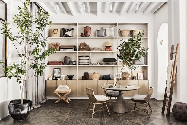 Bright, refined dining and display space with a round dining table and woven chairs set in front of extensive built-in open shelving and lower cabinetry. The room features white exposed ceiling beams, pale walls, dark patterned timber flooring, sheer curtains, potted trees, and layered neutral-toned decorative objects. A tall arched opening and leaning timber ladder add architectural and styling interest.