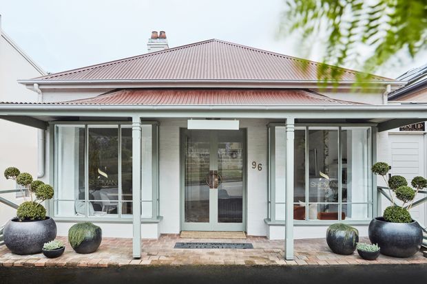 Front exterior view of a refined single-storey residence with a pale rendered facade, prominent hipped metal roof, covered veranda, glazed entry door, and symmetrical bay windows. Potted topiary plants line the paved entrance, with neighbouring buildings visible at the sides.