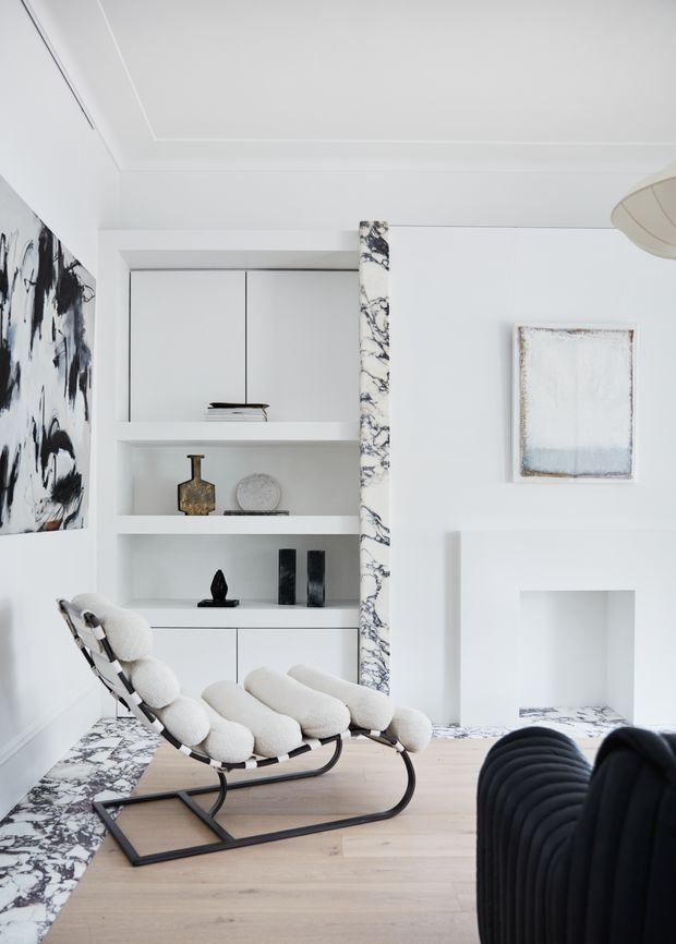 Bright, refined living room with white built-in shelving and cabinetry, pale timber flooring, dramatic marble-look veining used as a vertical trim and floor border, sculptural black-and-white lounge chair, black ribbed sofa, abstract artwork, and a recessed fireplace or display niche.