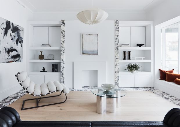 Bright, refined living room with white built-in shelving and cabinetry flanking a central recessed fireplace or display niche. Veined marble-look trim frames the joinery and continues along the floor perimeter. The room features pale timber flooring, a sculptural white pendant light, a black leather sofa in the foreground, a white-and-black lounge chair, a glass coffee table, contemporary artwork, and a window seat with rust-coloured cushions.