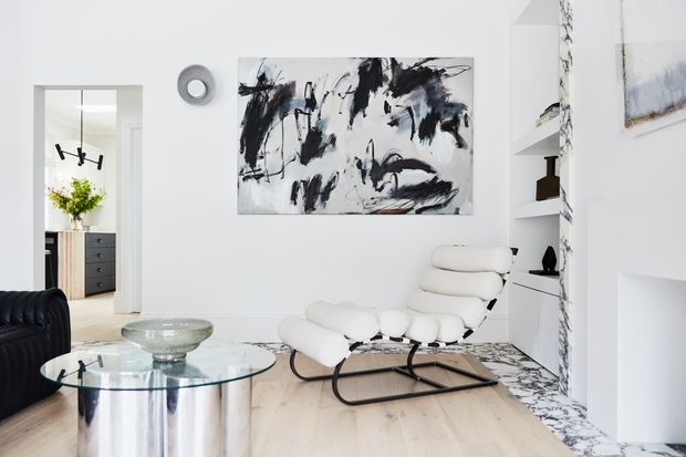 Bright, refined contemporary living room with white walls, pale timber flooring, a sculptural white lounge chair, black leather sofa, glass coffee table, built-in shelving, and bold black-and-white abstract artwork. A marble-clad fireplace or feature wall adds contrast, while a doorway reveals part of an adjacent room.