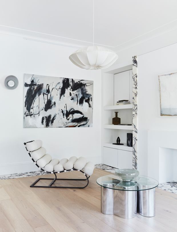 Bright, refined living room with white walls and decorative mouldings, pale timber flooring, a sculptural white pendant light, abstract black-and-white artwork, built-in open shelving, and a distinctive cream lounge chair beside a polished metal and glass coffee table. Marble-patterned accents appear around the shelving and fireplace area.