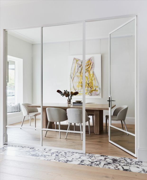 Bright, refined dining room viewed through white-framed glazed partition doors. The room features a long natural timber dining table, upholstered dining chairs, pale timber flooring, white walls and ceiling, and abstract yellow artwork. A marble-patterned threshold or flooring detail runs across the foreground, while a side window brings in abundant natural light.