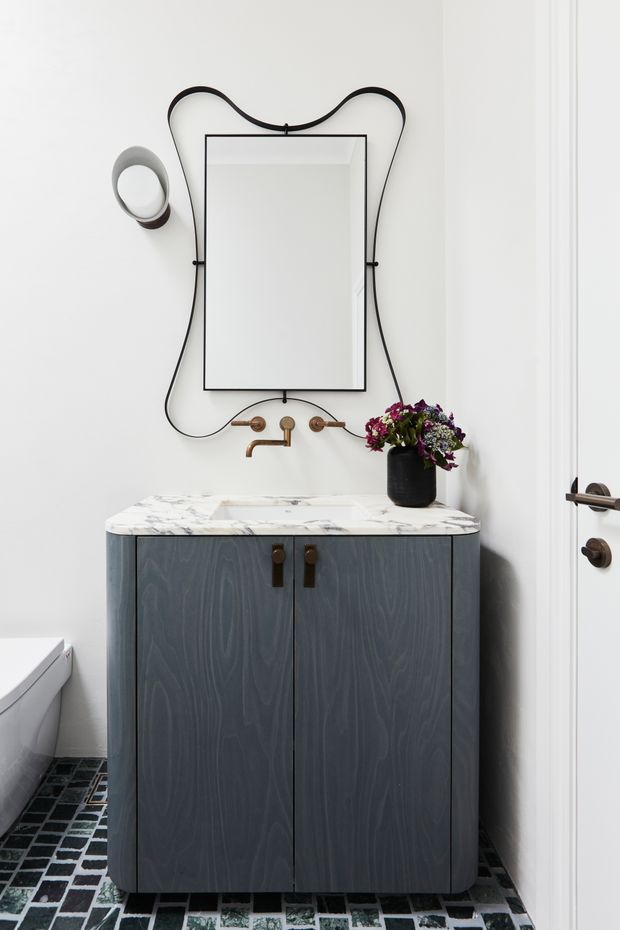 A compact, refined bathroom featuring a dark blue-grey timber vanity with a white marble benchtop, wall-mounted brass tapware, and a rectangular mirror framed by a distinctive looping black cord or trim. The room has white walls, a patterned dark geometric floor tile, contemporary wall lighting, and a floral arrangement on the vanity.