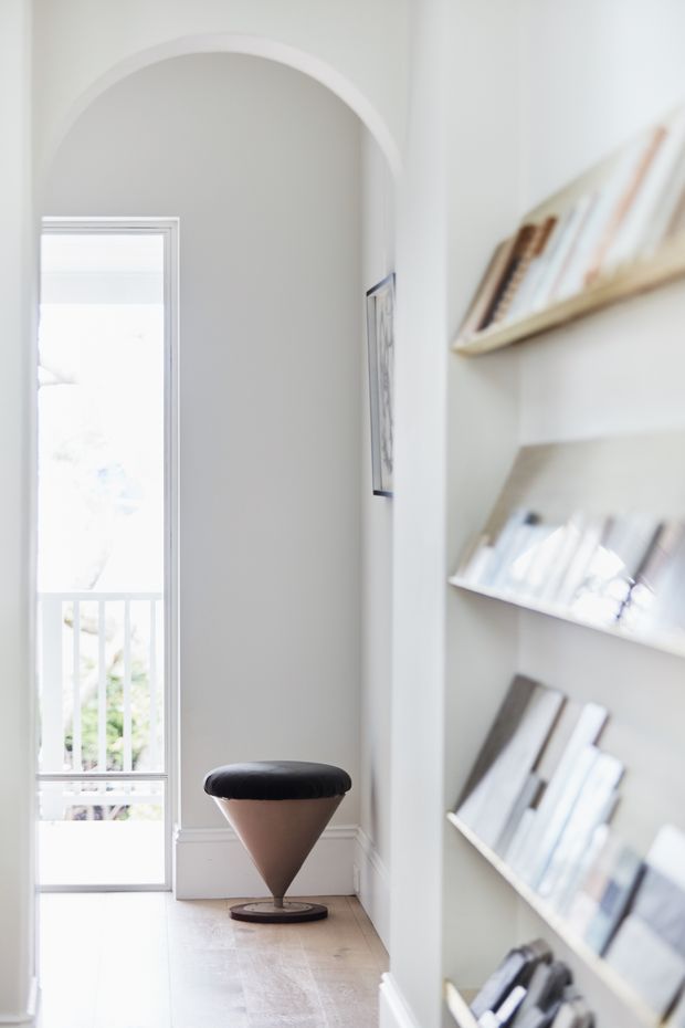 Bright, narrow interior hallway or landing with an arched wall opening, pale painted walls, light timber flooring, and a glazed door leading to a balcony or exterior. Angled built-in display shelves filled with books line the right wall, while a sculptural black-and-tan stool provides a focal point. The space has a refined, airy, minimalist-traditional character.