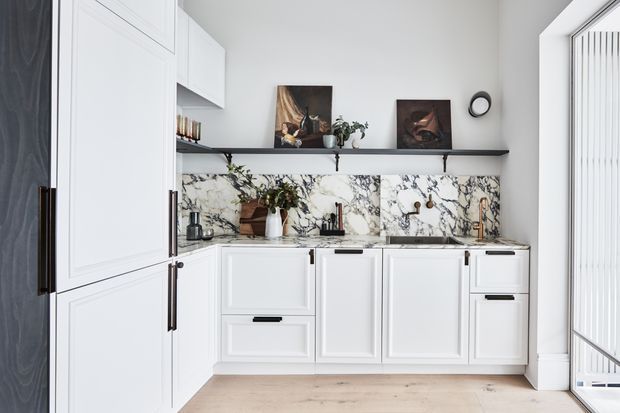 Bright, compact kitchen with white Shaker-style cabinetry, long dark hardware, open black metal shelving, and a dramatic veined marble backsplash. A tall dark timber pantry cabinet frames the left side, while a pale timber floor and vertical white window covering add contrast. The space is styled with small appliances, cookware, plants, and artwork.