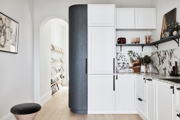 Bright, refined kitchen with white Shaker-style cabinetry, a dark curved timber pantry or feature cabinet, marble-look veined splashback, open black shelving, and pale timber flooring. An arched opening leads to a book-lined hallway, while a small upholstered stool and framed artwork add residential character.