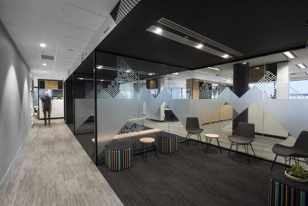 A contemporary commercial office interior showing a carpeted corridor beside a glazed meeting or breakout area. The space features black-framed glass partitions with frosted geometric privacy graphics, dark carpet, a black suspended ceiling canopy with integrated linear lighting, and casual seating arranged around small round tables. The palette is predominantly grey, black, and white with colourful striped cylindrical stools and planters providing accents.