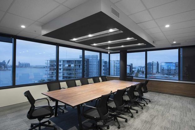 A contemporary commercial meeting or boardroom with a large timber conference table surrounded by black ergonomic office chairs. Floor-to-ceiling corner glazing provides expansive urban and waterfront views. The room features a suspended acoustic ceiling with recessed linear lighting, dark ceiling feature panels, black window frames, and a carpeted floor.