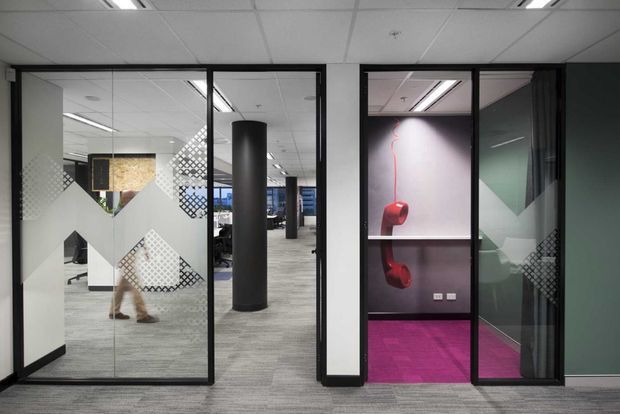 Interior view of a contemporary commercial office with glazed meeting rooms and partitions. Black-framed glass doors and geometric frosted privacy graphics define the foreground, while grey carpet tiles, white walls, a suspended acoustic-tile ceiling, and linear fluorescent lighting create a functional office environment. A vivid magenta carpeted room and display niche provide a strong accent colour, with workstations visible beyond.