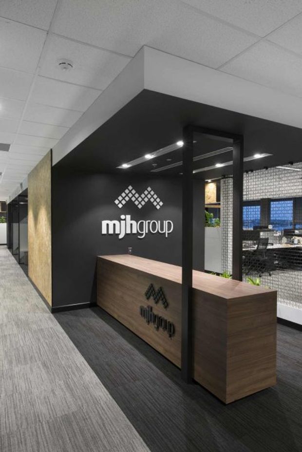 A contemporary commercial office reception area viewed from a corridor. A dark charcoal reception wall and canopy frame a timber-fronted reception desk with integrated branding. Behind the desk, a black metal mesh partition reveals workstations and blue-toned glazing or panels. The office has a suspended acoustic-tile ceiling, recessed linear lighting, carpet flooring, and a restrained black, grey, and timber palette.