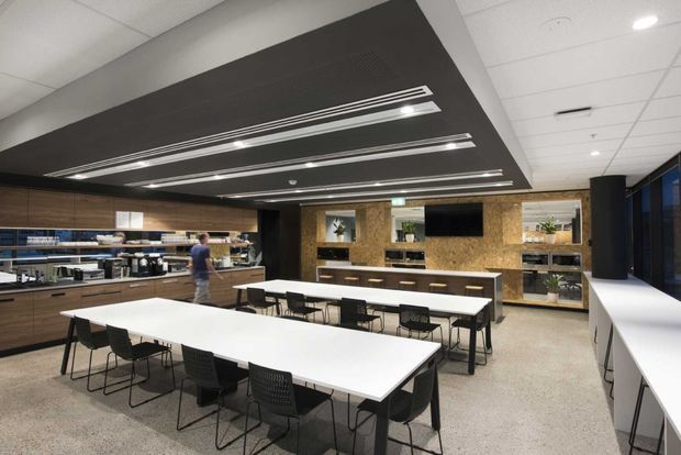 A contemporary commercial staff breakout and dining area with long white communal tables and black chairs. The room features dark timber cabinetry and shelving, integrated kitchenette facilities, a textured timber feature wall with recessed display niches and a wall-mounted screen, and a large dark ceiling bulkhead containing linear recessed lighting. A long white counter runs along the glazed right side.