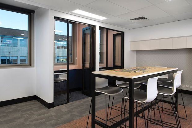 A contemporary commercial meeting or collaboration room with a long high table surrounded by white and grey bar stools. The room has glazed black-framed partitions and windows, pale built-in wall cabinetry, a patterned grey backsplash or wall surface, carpeted flooring, and a suspended acoustic tile ceiling with recessed lighting. Adjacent office or building areas are visible through the glazing.