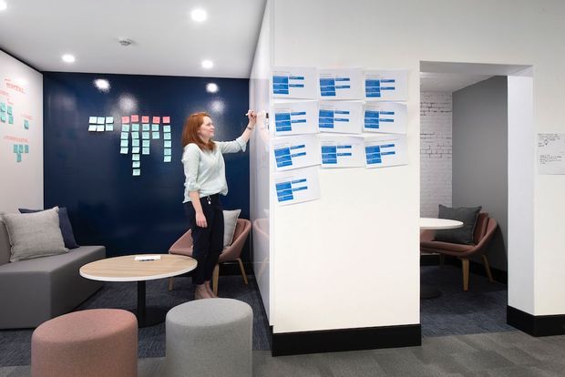 A contemporary collaborative office breakout area with a dark navy feature wall covered in sticky notes, a white planning board displaying blue-and-white cards, and a small meeting nook visible through an opening. The space includes a grey sofa, upholstered lounge chairs and stools, a round light-wood coffee table, grey carpet flooring, recessed ceiling lights, and black skirting.