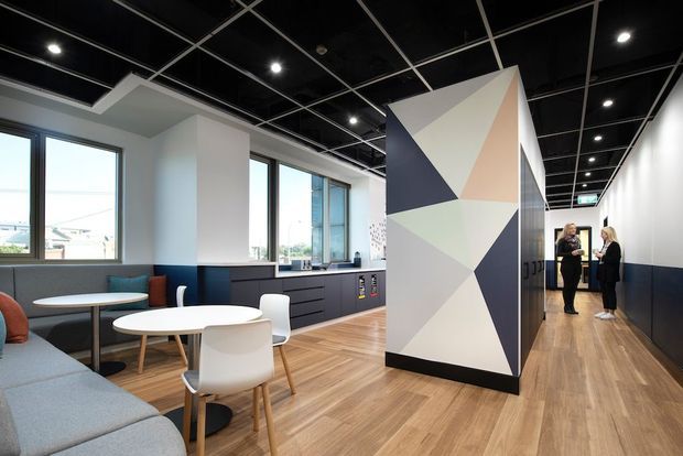 A contemporary commercial interior with an open breakout lounge and kitchenette or staff amenity area. The space features pale timber-look flooring, white walls, dark navy lower wall panelling and cabinetry, large windows, and a black exposed grid ceiling with recessed spotlights. A tall geometric abstract partition or feature wall divides the seating area from a corridor, with small round tables, white chairs, and a grey upholstered banquette. The corridor extends toward a doorway where two people are visible.