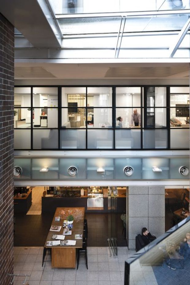 A vertical view into a multi-level contemporary commercial interior, likely an office, hospitality, or retail atrium. The image shows black-framed glazed partitions on an upper level, exposed brick and concrete surfaces, a large overhead skylight, mechanical ventilation grilles, and a lower-level café or communal area with a long timber table and dark flooring. People are visible in the background, while a glazed balustrade and stair edge appear in the foreground.