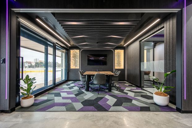 A sleek contemporary meeting room with floor-to-ceiling glazing, dark ribbed wall panels, a dramatic linear slatted ceiling, integrated strip lighting, and a central conference table with office chairs. The patterned geometric carpet uses grey, purple, and muted green tones, while potted plants and a wall-mounted screen complete the corporate interior.
