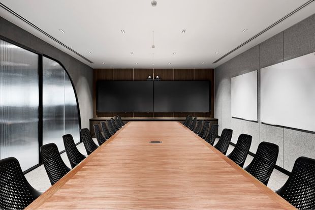 A sleek contemporary boardroom with a long timber conference table, black perforated chairs, dark wood wall panelling, acoustic wall panels, integrated display screens, and curved black-framed glazing. The room has a clean, minimalist corporate aesthetic with recessed linear lighting and a neutral grey, black, and warm timber palette.