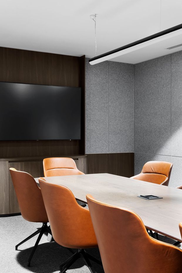A refined contemporary meeting room with a large light timber conference table surrounded by tan leather swivel chairs. Dark timber wall panelling and built-in cabinetry frame a wall-mounted black display, while grey acoustic wall panels provide texture and sound control. A linear suspended light and concealed ceiling air-conditioning complete the professional commercial fit-out.