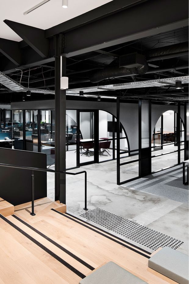 A contemporary commercial office interior viewed from a stepped entry or mezzanine. The space features exposed black structural steel beams and services, black-framed glazed meeting rooms with distinctive arched partitions, open workstations, polished concrete flooring, pale timber steps with black contrasting inlays, black metal balustrades, and linear recessed and track lighting. The palette is predominantly black, white, grey, and light timber, creating an industrial yet refined workplace atmosphere.