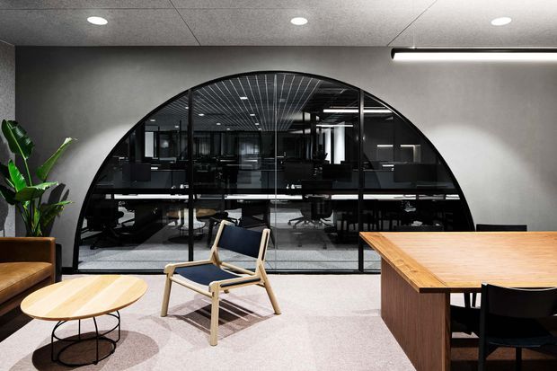 A contemporary commercial office interior with a dramatic arched black-framed glazed partition overlooking an open-plan workspace. The foreground includes a timber meeting table, upholstered lounge chair, leather seating, and an oval coffee table. Dark grey walls, a textured acoustic ceiling with recessed spotlights, linear pendant lighting, and neutral carpet create a refined, professional atmosphere.