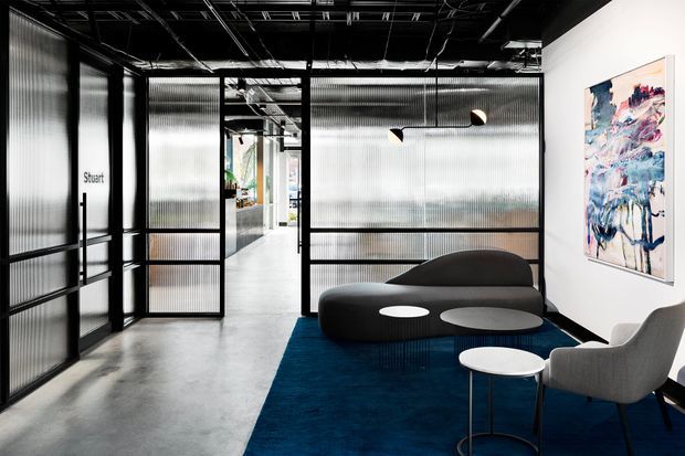 Contemporary commercial office lounge and circulation area with translucent corrugated glass partitions in black metal frames, exposed black ceiling services, polished concrete flooring, a deep blue rug, sculptural dark seating, a light grey armchair, and a small round table. Abstract artwork adds colour to the otherwise monochrome interior.