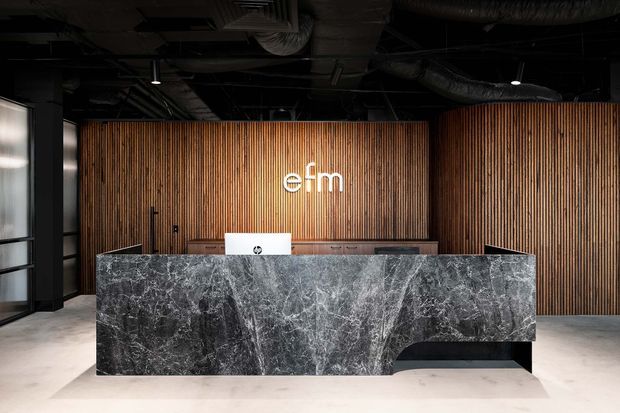 A dark, sophisticated commercial reception area with a large black-and-grey stone reception desk in the foreground. The desk sits against a full-height vertical timber-slat feature wall with an illuminated logo, while exposed black ceiling services, ducts and lighting contribute an industrial character. Large glazed panels are visible at the left.