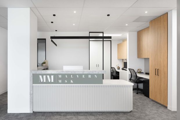 Bright contemporary office reception area with a white fluted-front desk, grey carpet, white walls and ceiling tiles, black-framed glass partitions, and warm timber built-in cabinetry and workstations. The space includes visible office chairs and desks behind the reception counter.
