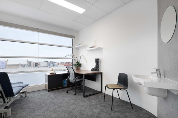 Bright contemporary office consultation room with a large street-facing window, two workstations, visitor chair, grey carpet, white walls, suspended acoustic ceiling, and a compact wall-mounted basin against a grey feature wall.