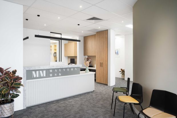 Bright contemporary commercial reception area with a white fluted reception desk, grey carpet, timber veneer cabinetry and wall panels, black linear pendant lighting, and a small waiting area with modern chairs. The space has a clean, professional medical or office-like fit-out with indoor plants and a suspended acoustic tile ceiling.