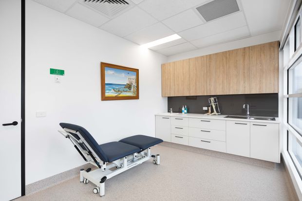 Bright, clean clinical treatment room with an adjustable examination chair, white built-in cabinetry, a dark benchtop and splashback, timber-look overhead cupboards, large windows, and a suspended acoustic-tile ceiling. A coastal artwork and sink area add minor visual detail.