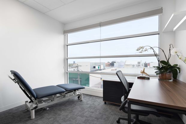 Bright, compact professional consultation or treatment room with a reclining examination chair, office desk, ergonomic chair, orchid, broad windows, and a city-facing view. The space has white walls, a suspended acoustic ceiling, grey carpet, and dark wood office furniture.