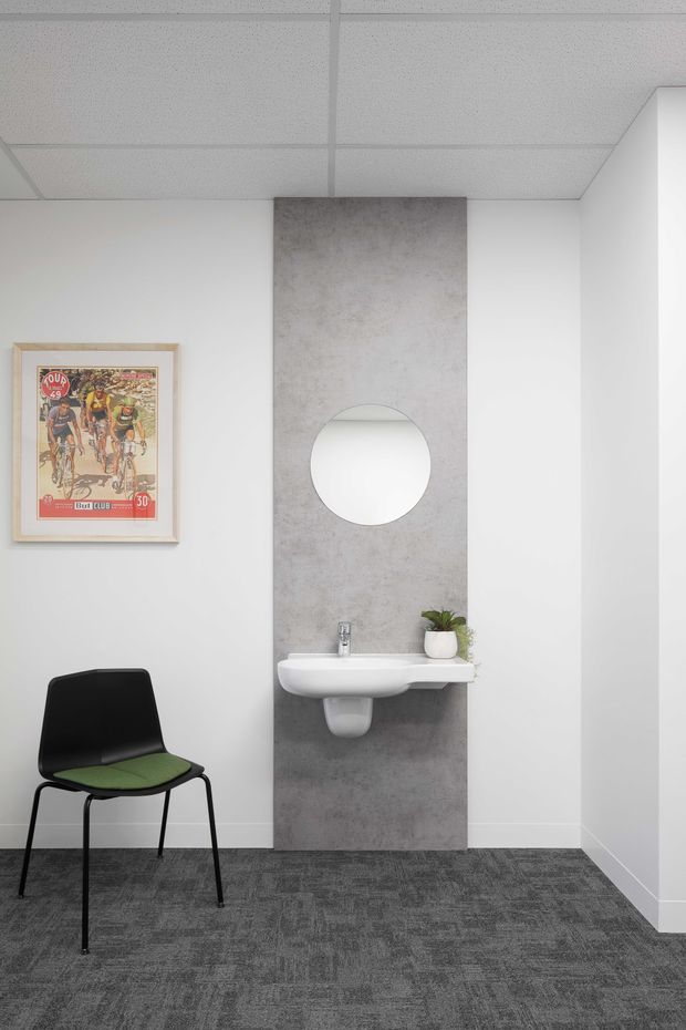 A compact, minimalist bathroom or washroom with a wall-mounted white basin, circular mirror, and a tall grey concrete-look feature panel. The room has white walls, a suspended acoustic tile ceiling, dark grey carpet flooring, and a black chair with a green seat cushion. A framed cycling poster and small potted plant add limited decorative detail.
