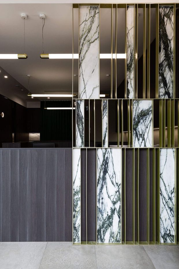 A sophisticated commercial reception or hospitality interior featuring a dramatic decorative partition of vertically arranged white veined marble panels, dark timber or laminate panels, and slender brass-toned metal framing. Behind it is a dark reception counter with grey woodgrain cladding, black walls, and linear pendant lighting. Large pale stone or concrete-look floor tiles and a restrained dark palette create a refined, luxurious atmosphere.