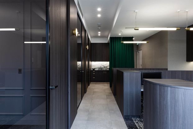 A refined contemporary commercial interior with a narrow tiled circulation space, dark timber wall panelling and doors, glazed office partitions, and a dark reception or workstation counter in the foreground. Linear pendant lights and recessed ceiling lights illuminate the space, with a deep green curtain or partition adding a restrained accent.