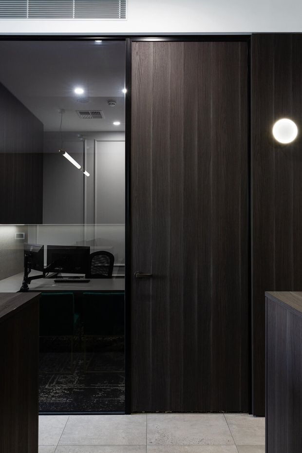 A dark, refined interior featuring full-height dark timber veneer doors and cabinetry, a glass-partitioned home office visible beyond, and a pale tiled floor. Recessed ceiling lighting and a circular wall light create a moody, contemporary atmosphere.