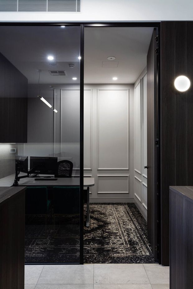 A compact, refined home office viewed through a dark-framed glass doorway. The office contains a desk with computer equipment, dark built-in cabinetry, a black patterned rug and upholstered seating. The adjacent hallway features decorative white wall panelling, recessed ceiling lights, a patterned runner, and dark timber joinery. Large pale floor tiles are visible in the foreground.