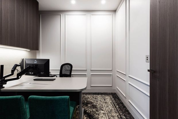 A compact, refined home office with a white desk, computer workstation, ergonomic chair, and dark green upholstered visitor chairs. The room features dark wood built-in cabinetry, white decorative wall panelling and trim, recessed ceiling lights, and a patterned dark rug.