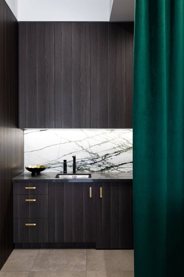 Compact, highly styled bathroom vanity with dark vertically grained timber cabinetry, a pale stone benchtop and dramatic white marble splashback with dark veining. A black wall-mounted faucet and basin sit beneath upper cabinetry, with brass-gold handles and a deep emerald green curtain or screen at right. Large neutral stone-look floor tiles complete the space.