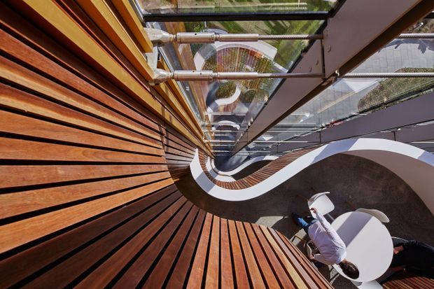 Abstract architectural view of a contemporary multi-level interior atrium or stairwell, photographed from above with strong geometric distortion. Warm timber slatted wall and floor surfaces curve around a central void, contrasted with sweeping white sculptural balustrades, dark structural surfaces, and extensive overhead glazing. A small seating area with white chairs and a round table is visible below.