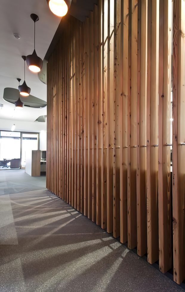 A contemporary interior corridor or open-plan office zone featuring a full-height vertical timber batten screen along the right side. Warm natural timber slats filter strong sunlight onto a dark carpeted floor, while the left side opens toward a bright workspace with desks, glazing, and sculptural pendant lights. The space has a refined, warm commercial interior character with strong linear shadows and detailing.