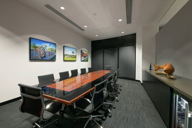 A contemporary commercial meeting or boardroom with a long glossy timber-and-dark-surface conference table surrounded by black wheeled office chairs. White walls display framed artwork, while dark full-height cabinetry or sliding panels occupy the rear wall. Recessed ceiling lights and linear air-conditioning grilles provide even illumination, and a built-in side counter includes decorative objects and a partially visible under-counter beverage refrigerator. The room has a polished, professional atmosphere with strong black, white, and warm timber contrasts.